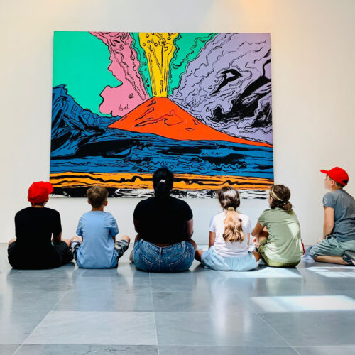 Foto di gruppo di bambini e una educatrice che guardano un'opera d'arte.